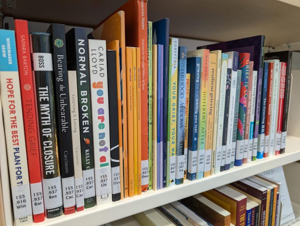 A row of library books about grief sits on an antique white metal shelf.