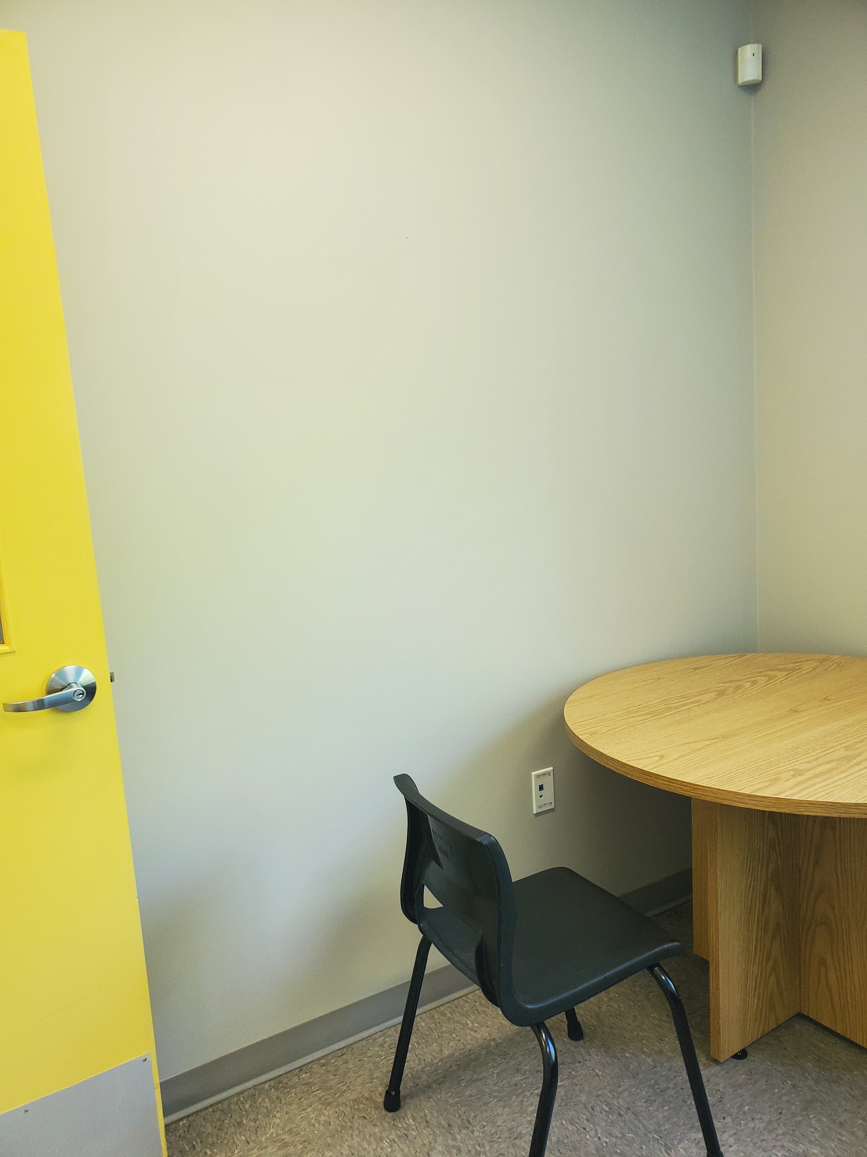 A yellow door opens to a blank beige wall. In the corner is a round wooden table and a black chair.