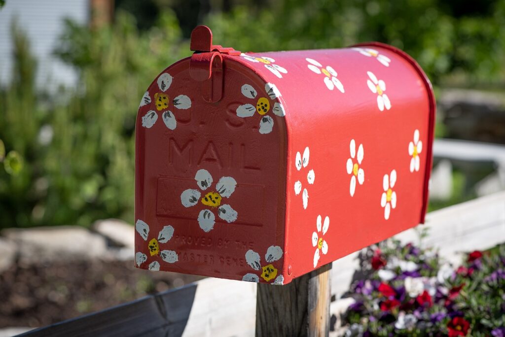 A red mailbox with yellow and white daisies painted on it. It sits on a fence rail, with blurred flowers and trees behind it.